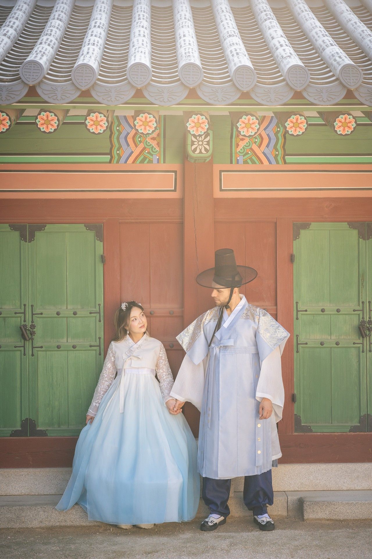 A couple in traditional Korean hanbok clothing sits on a wooden porch of a historic building.