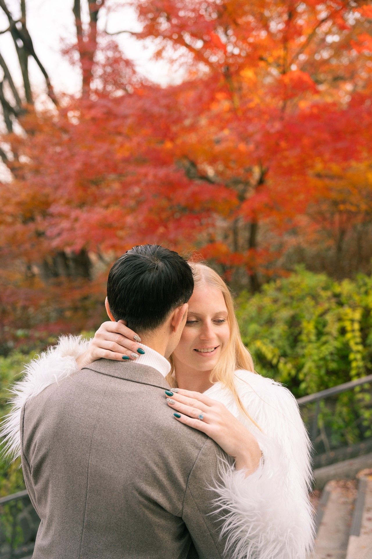 A couple embraces outdoors surrounded by vibrant red autumn maple trees in a romantic fall setting.