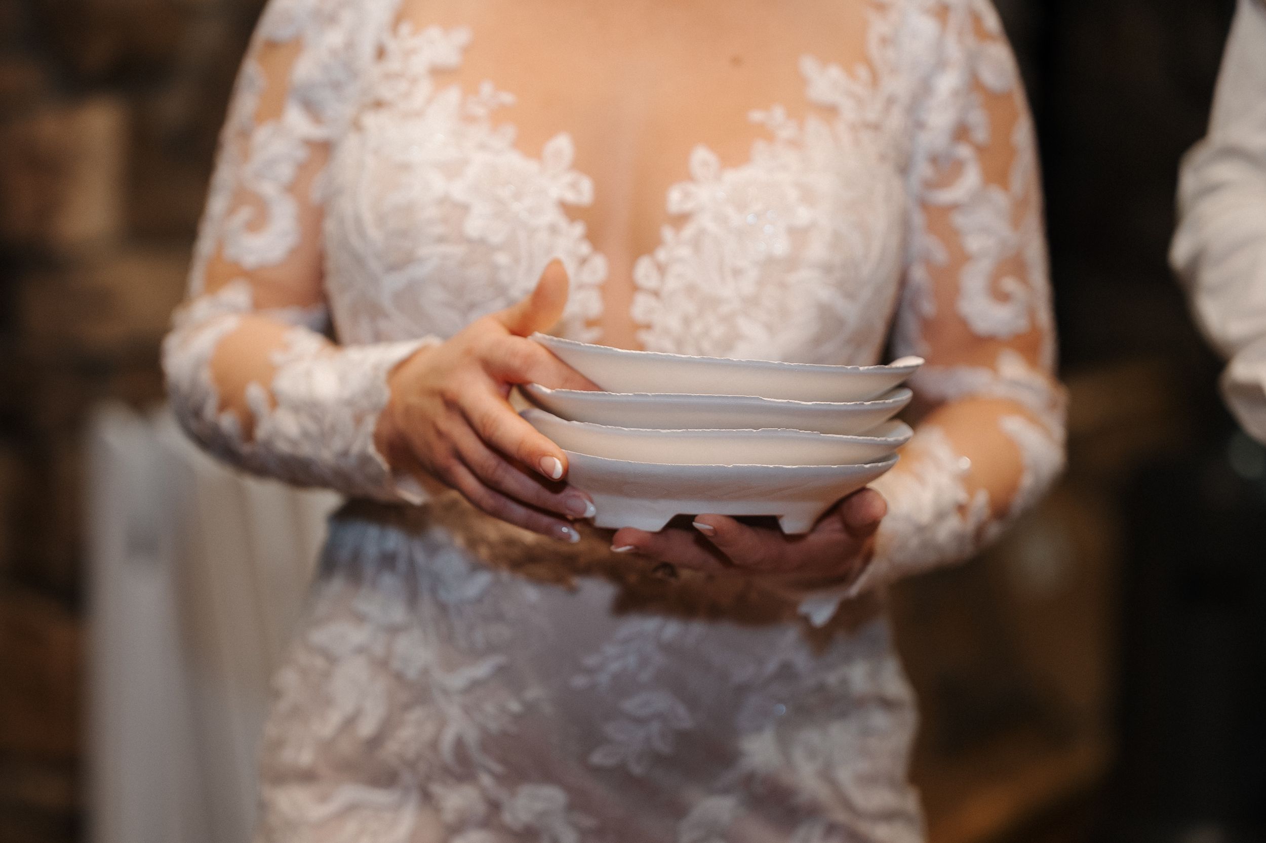 A person in a white lace wedding dress holds stacked white dinner plates in an elegant setting.