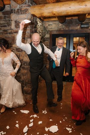 Wedding party members dance and stomp on napkins during a fun reception game at a rustic log cabin venue.