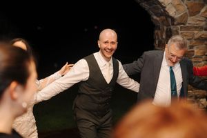 Groomsmen in formal attire laugh and celebrate together at a wedding reception in front of a stone wall.