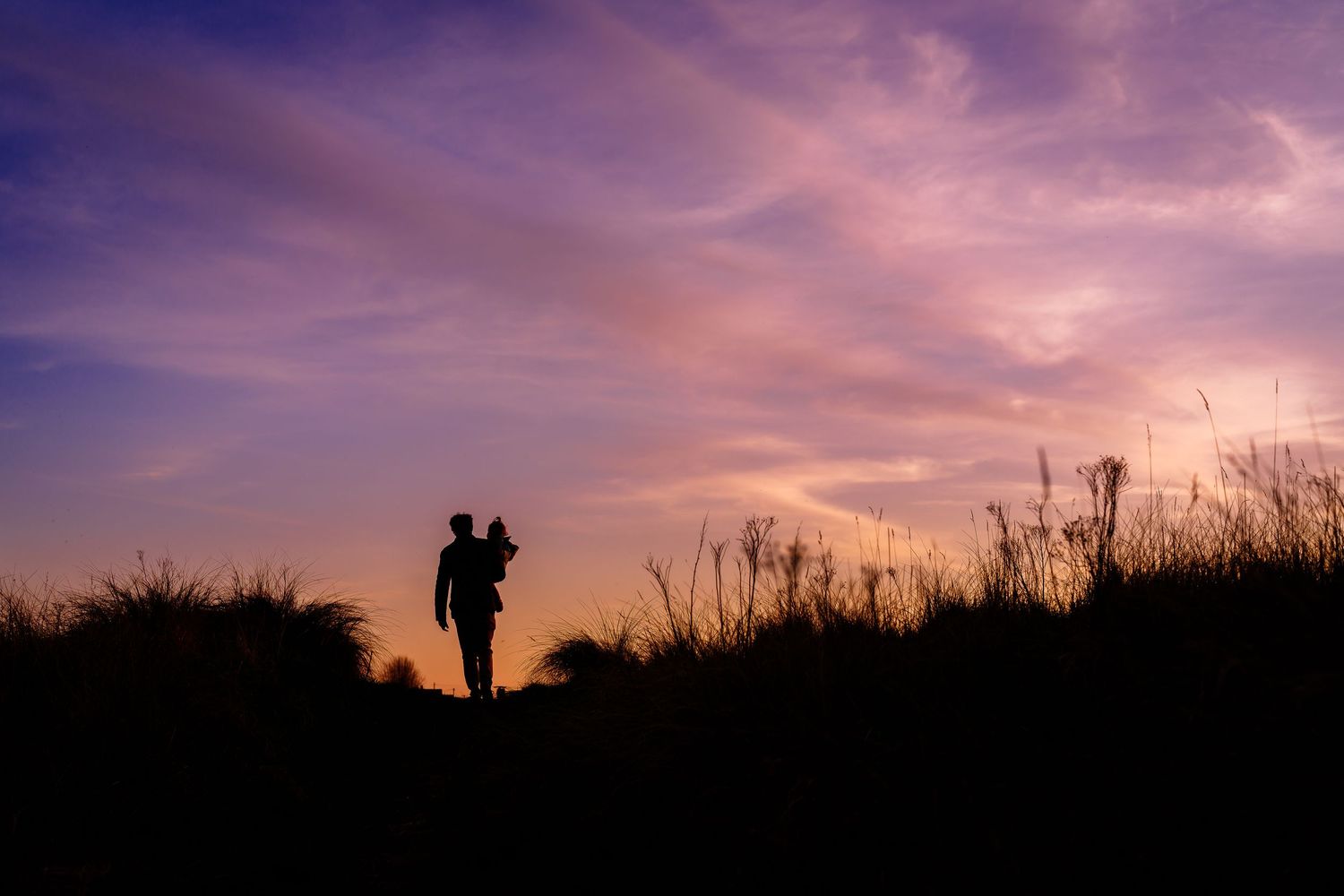 Silhouettes of people against a dramatic purple sunset sky with tall grass in foreground.