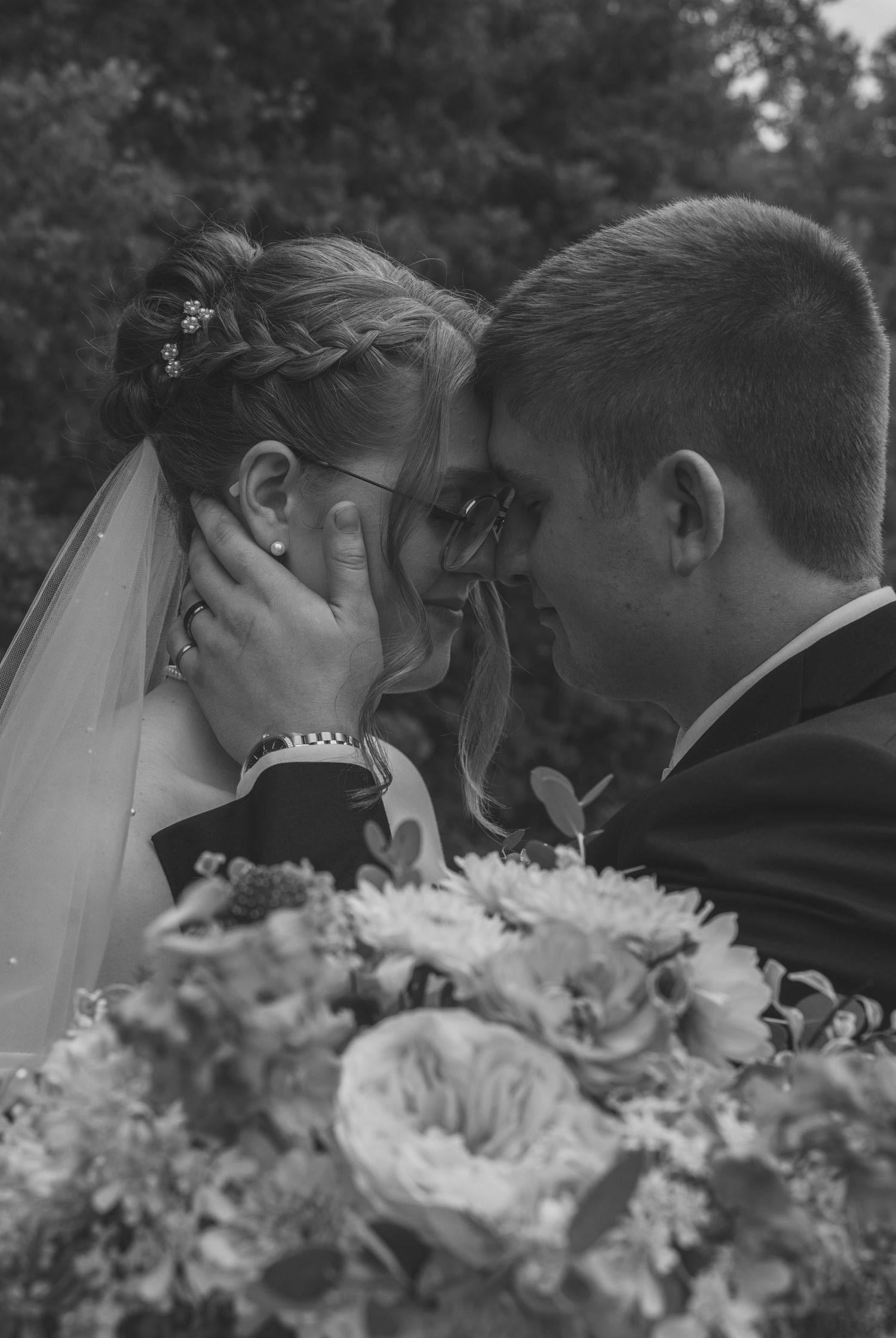 A romantic wedding couple shares an intimate kiss behind a bouquet of white flowers in black and white photography.