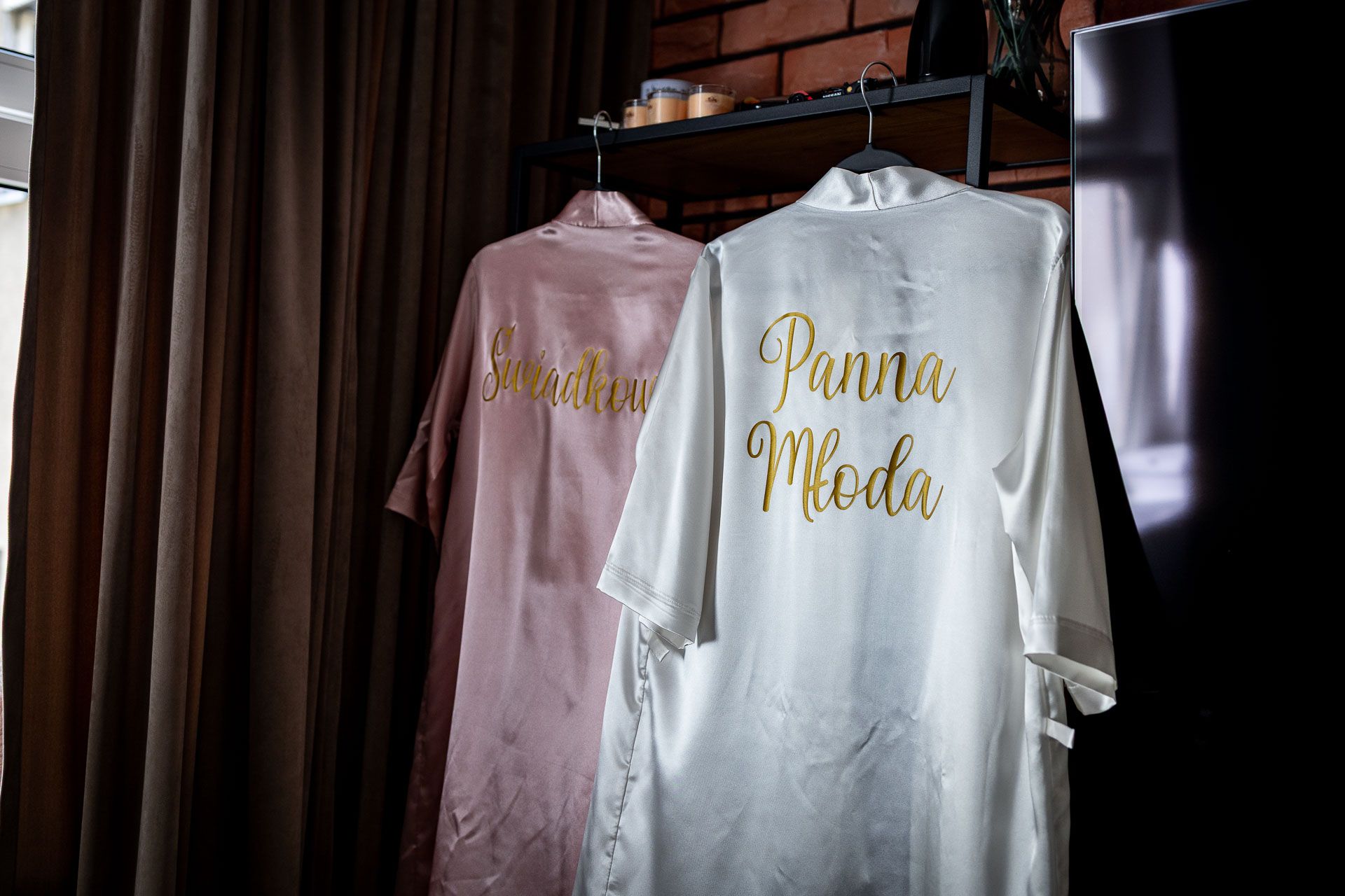 Pink satin robes with gold text hanging on wooden hangers against a brick wall.