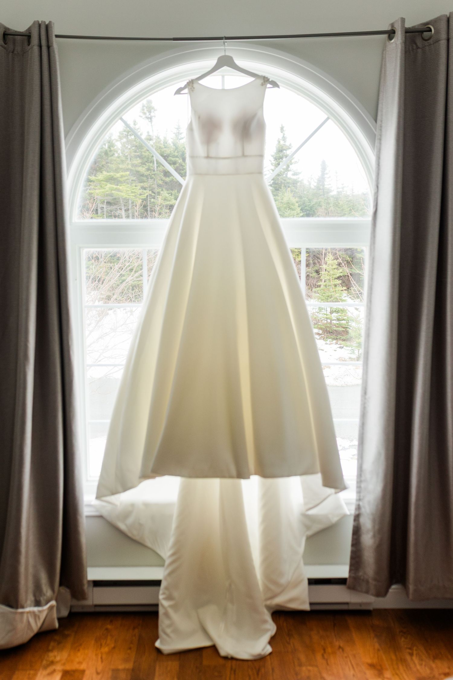 White wedding dress hanging in front of arched windows with dark curtains on a hardwood floor.