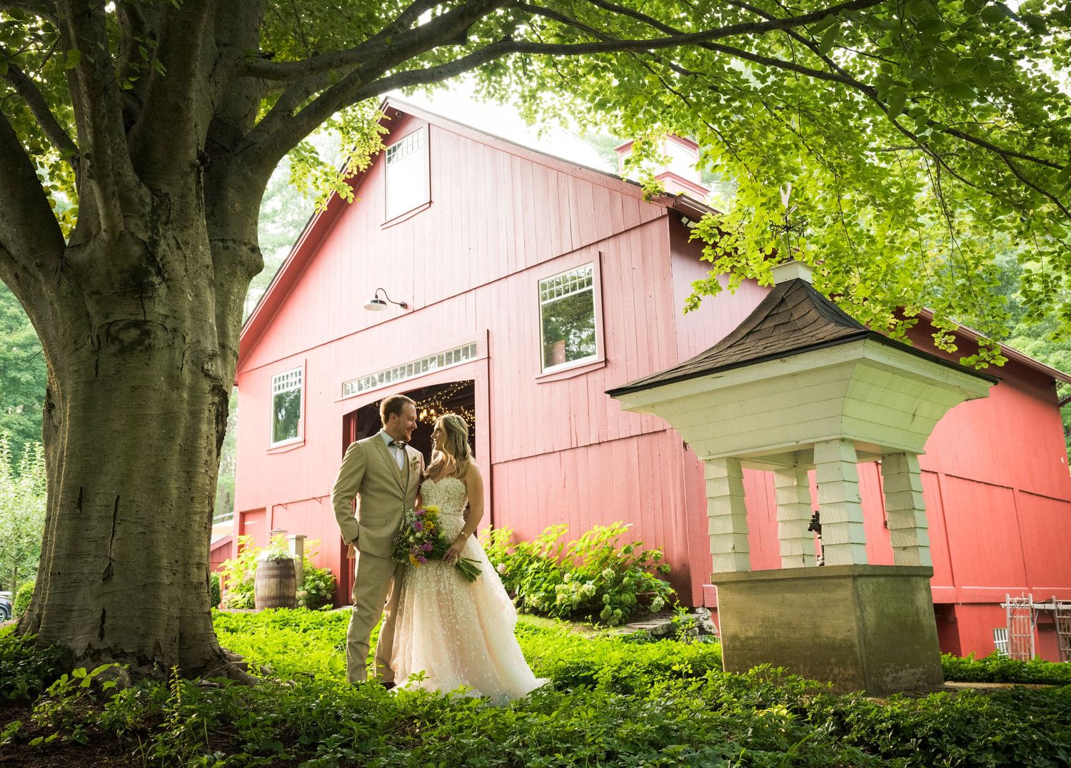 A couple stands together in front of a charming pink barn surrounded by lush green trees on their wedding day.