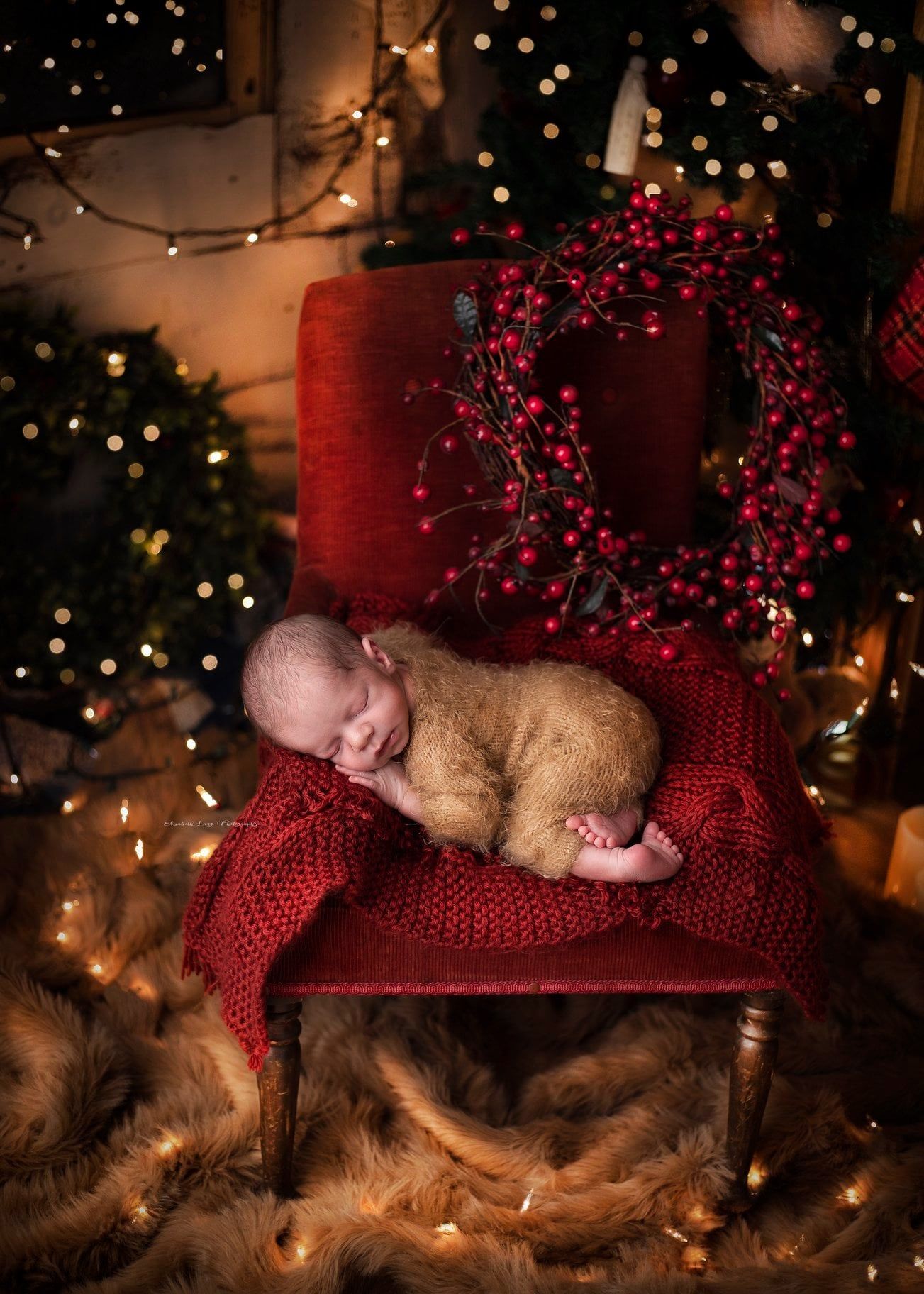 Sleeping baby wrapped in red knit blanket on brown fur with twinkling Christmas lights in background.