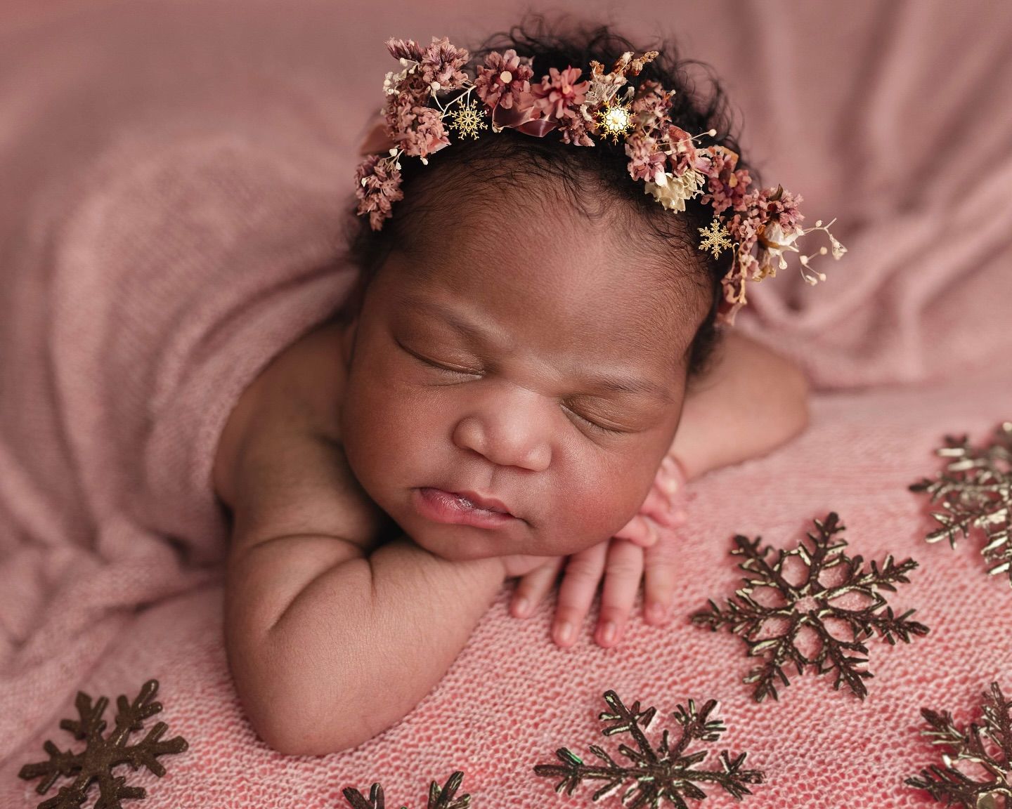 Sleeping newborn wearing delicate floral crown rests peacefully on pink blanket with snowflake pattern.