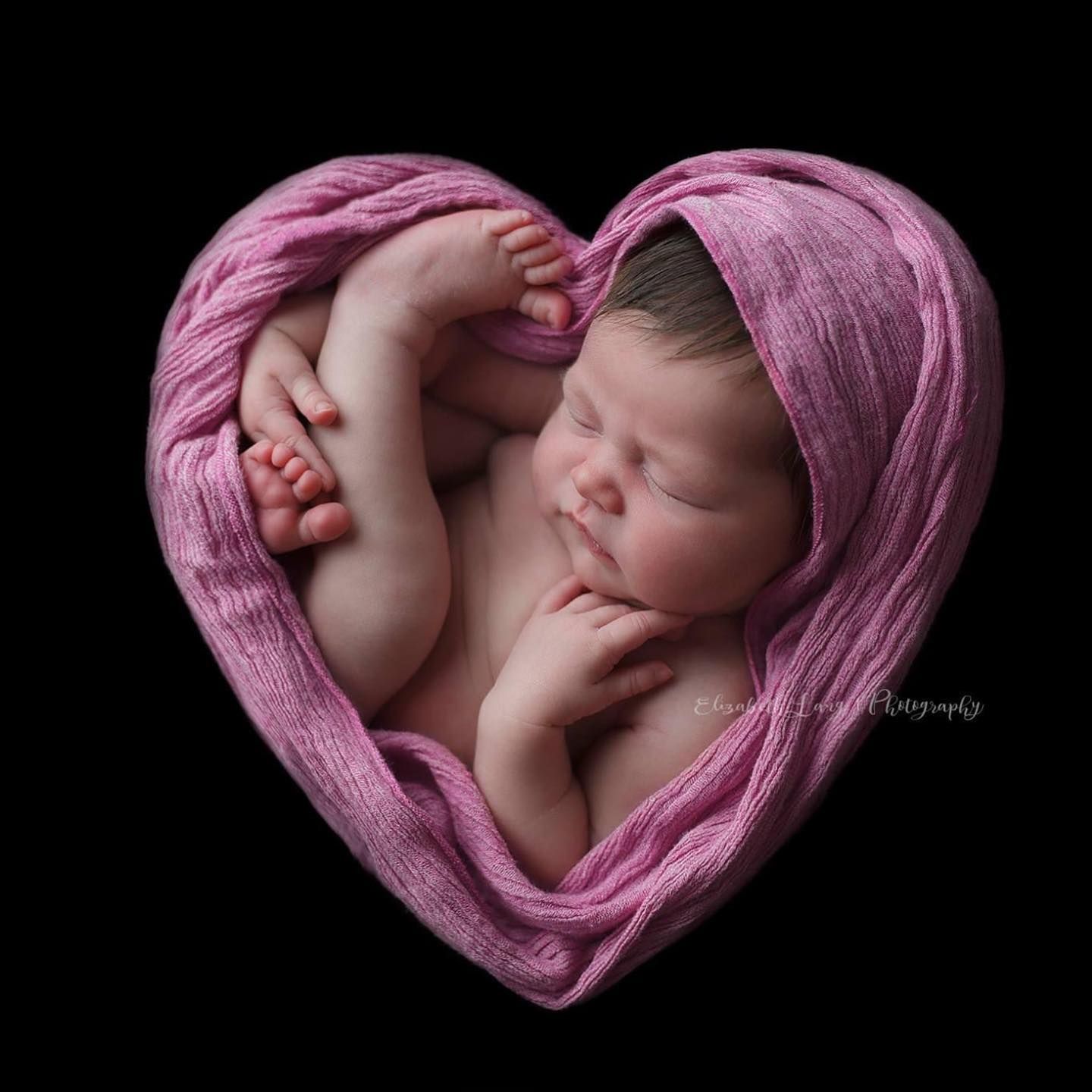 Newborn baby nestled in soft pink fabric arranged in heart shapes against black background creating artistic portraits.