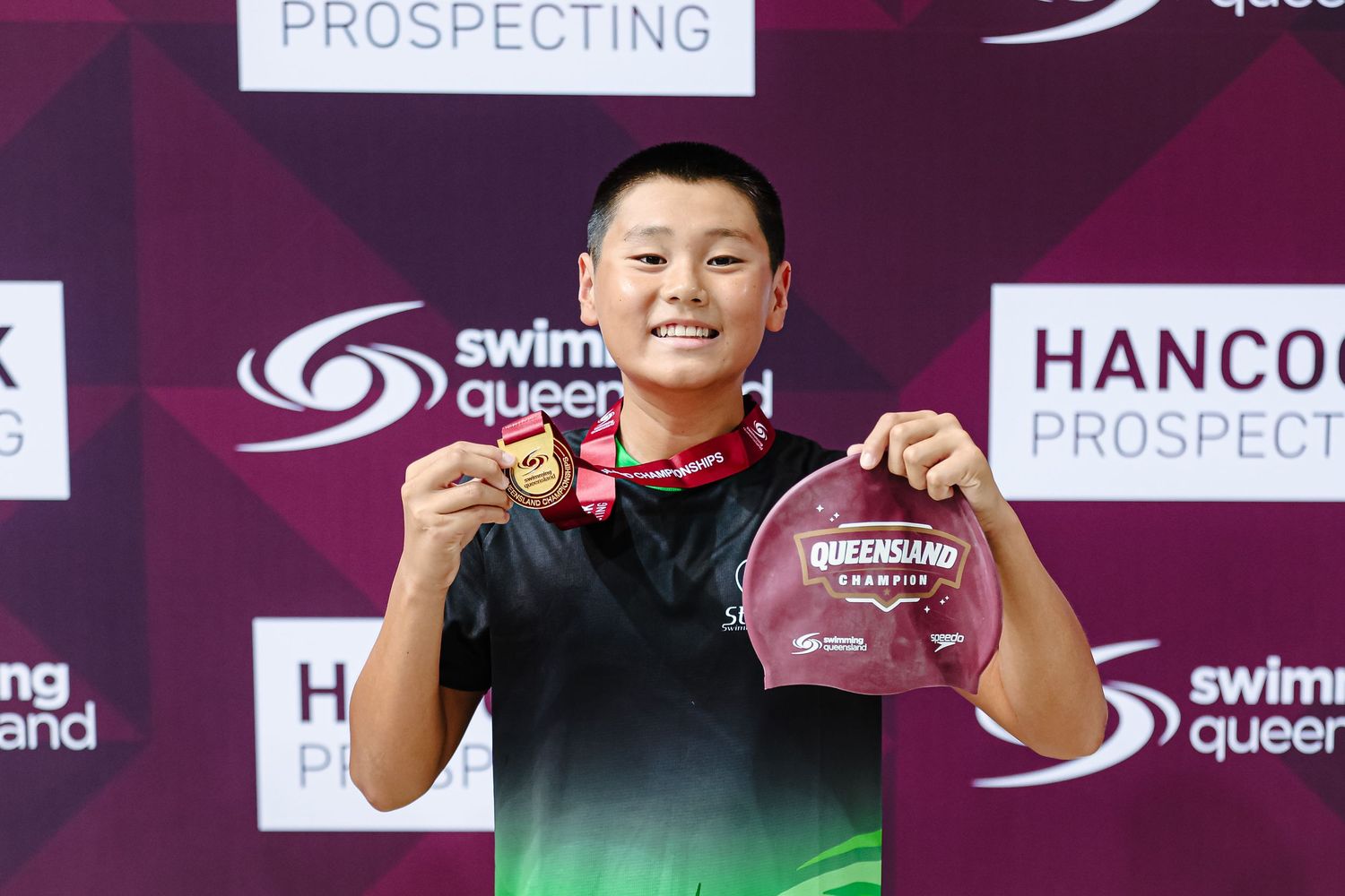 Two athletes stand on a podium displaying medals at a swimming competition with Hancock sponsorship banners in background.