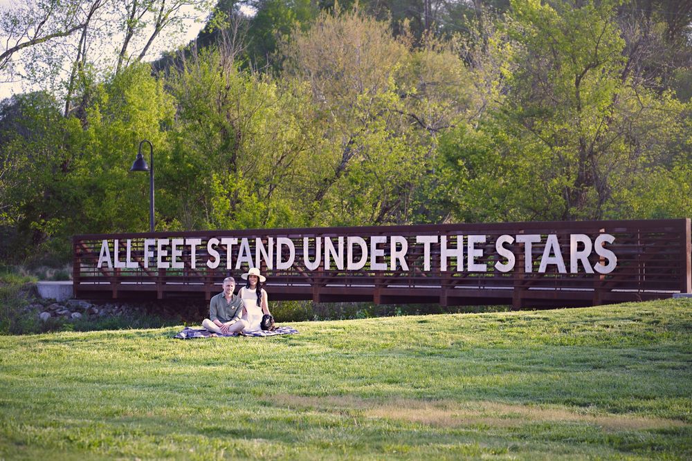 Scenic panoramic view of a green field with a large sign reading 'All Feet Stand Under The Stars' against trees.