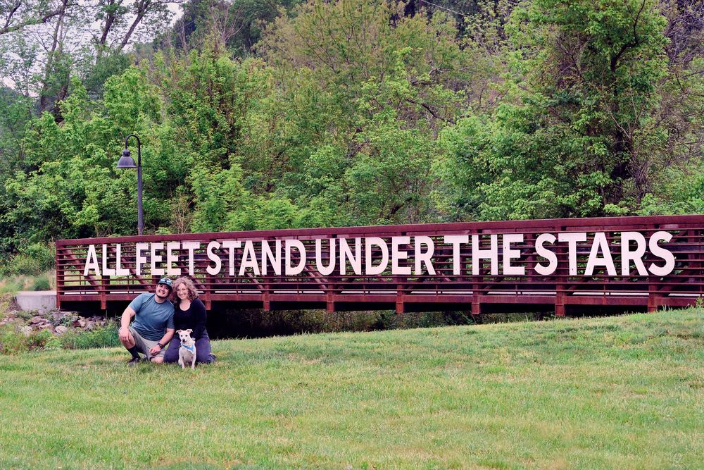 People enjoying a peaceful outdoor setting with a large sign reading 'All Feet Stand Under The Stars' in a grassy field.