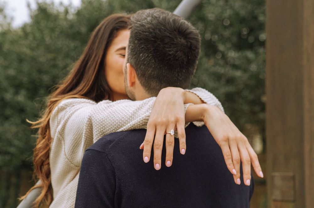 A couple embraces in a romantic moment outdoors with sunlight filtering through trees in the background.