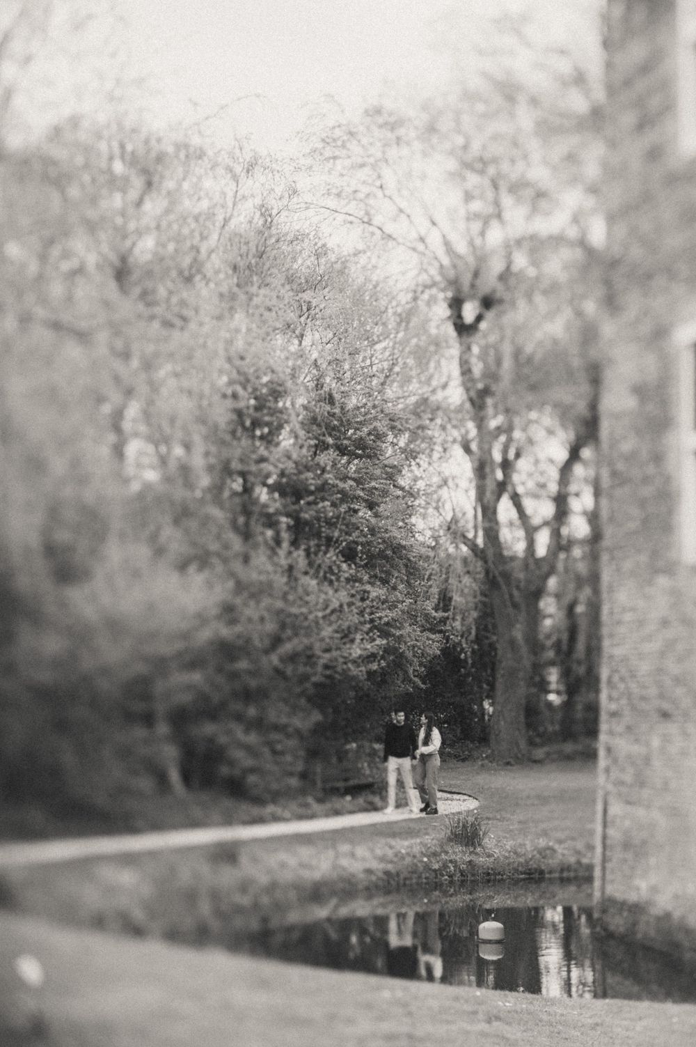 A black and white vintage photograph shows people walking along a tranquil canal path bordered by trees and foliage.