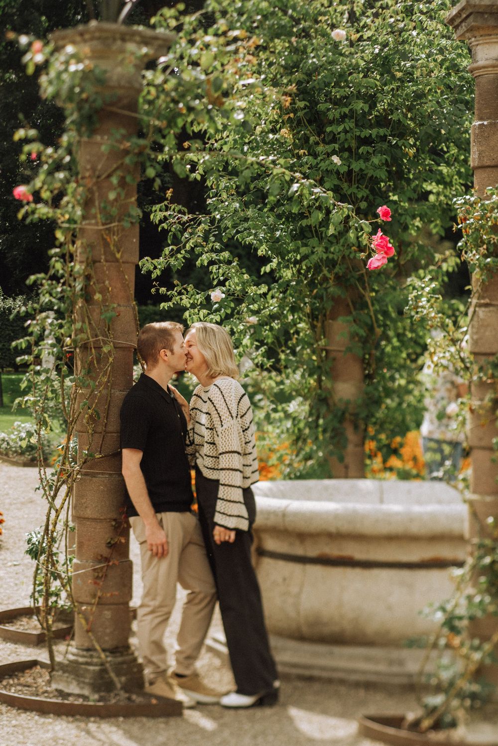 A couple shares a romantic moment in a garden setting with climbing roses and stone pillars.