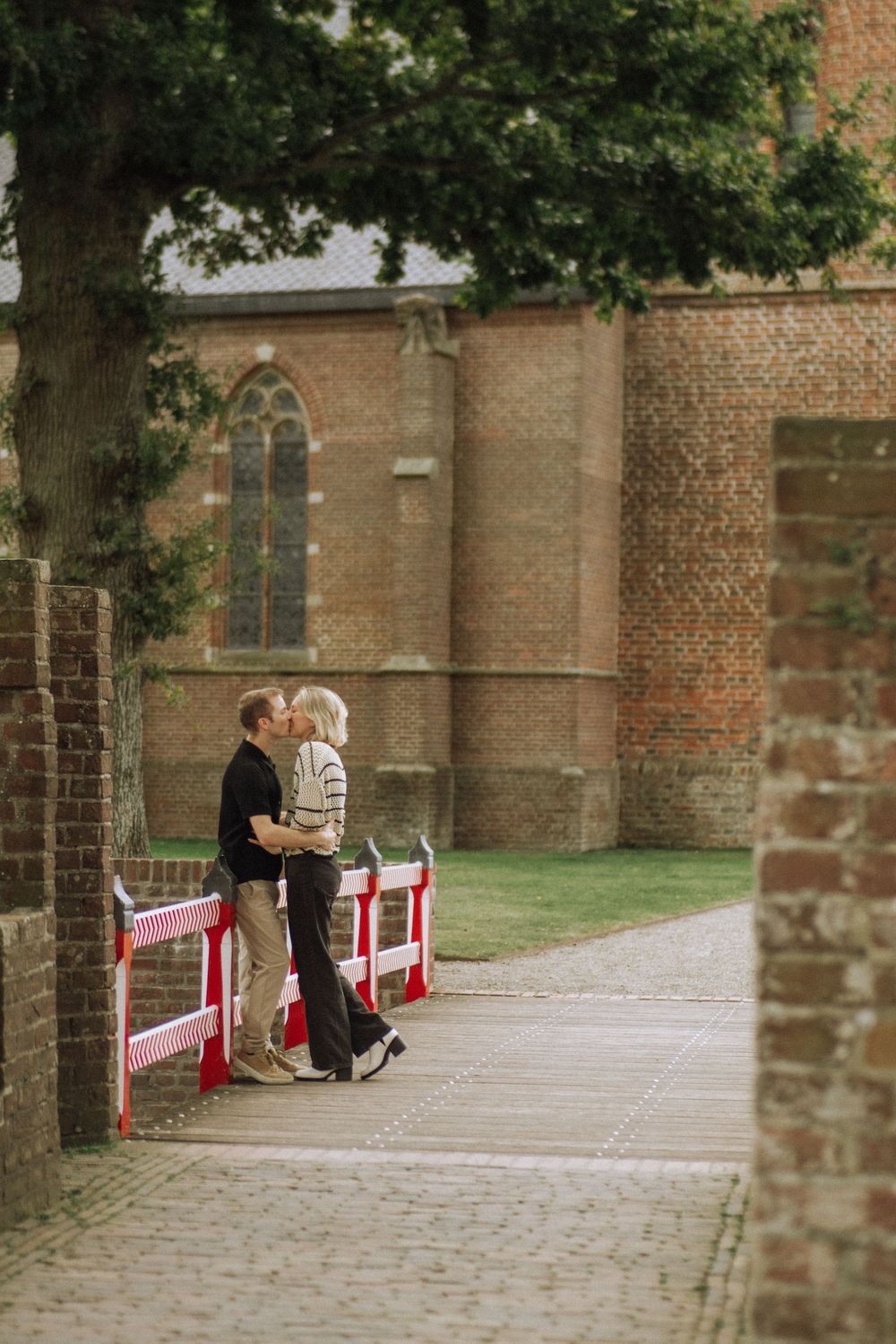 A romantic proposal scene unfolds on a cobblestone path beside a historic brick church wall.