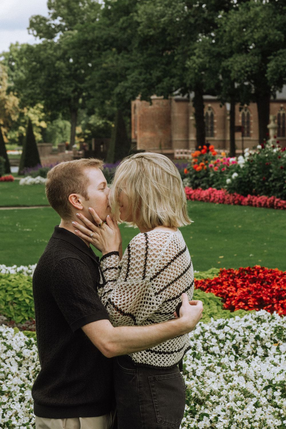 A couple shares romantic kisses in a garden with red and white flowers blooming in the background.