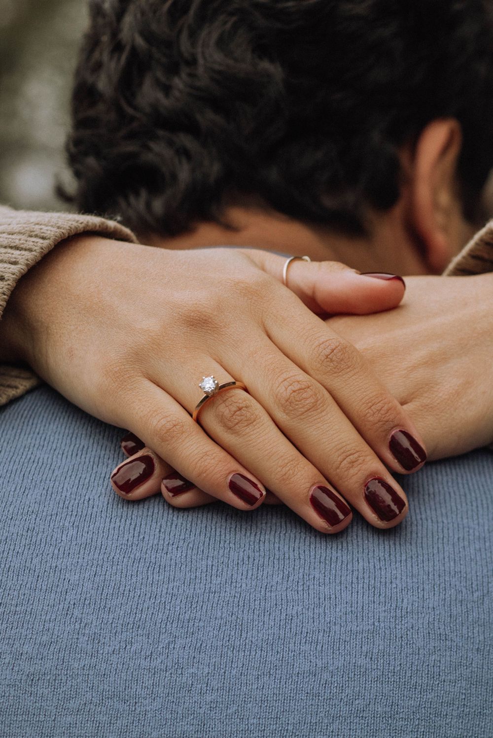 Close-up of a hand wearing burgundy nail polish and a sparkling engagement ring resting on grey fabric.
