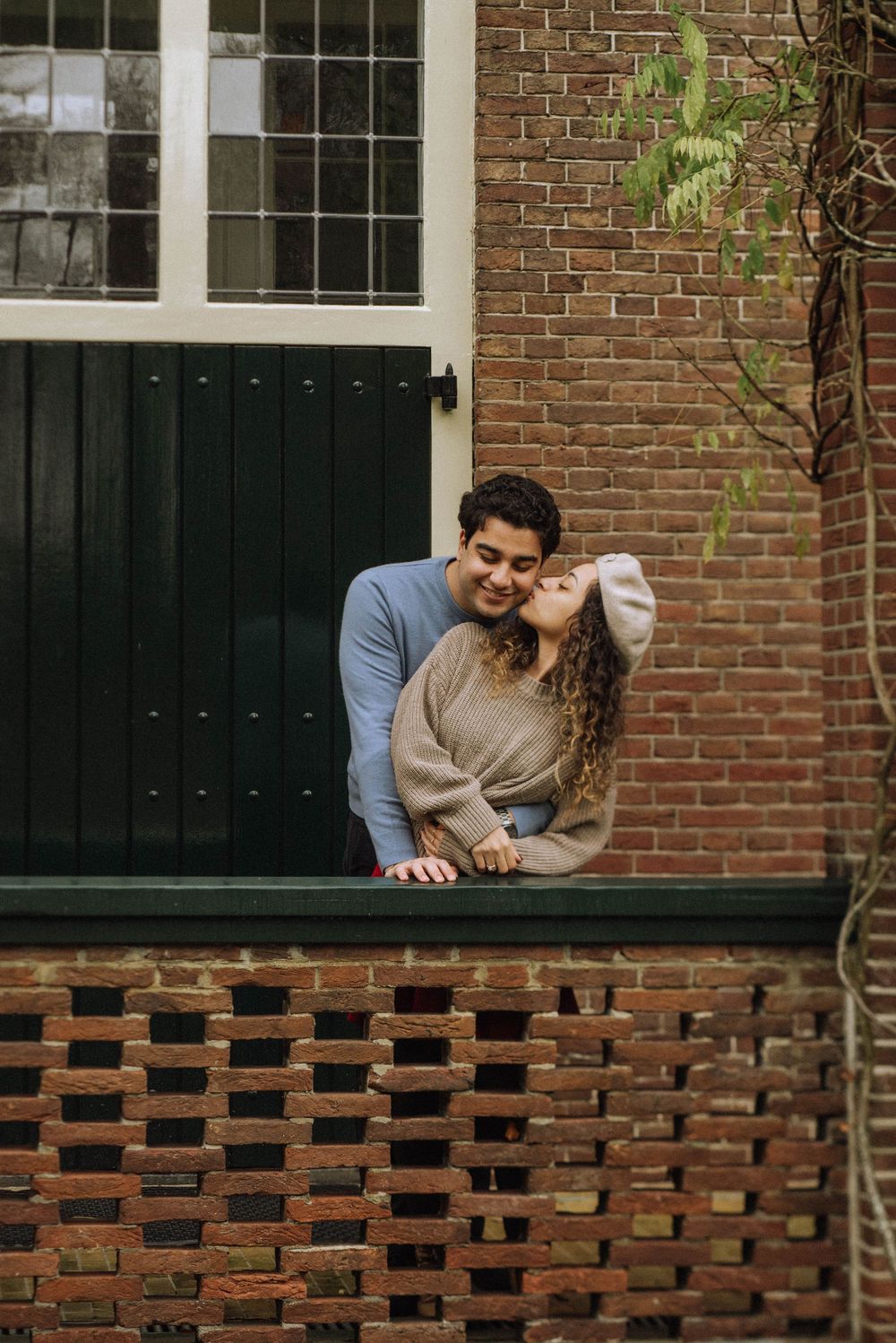 A couple shares a tender moment while sitting on a rustic brick wall outside a building with white-framed windows.