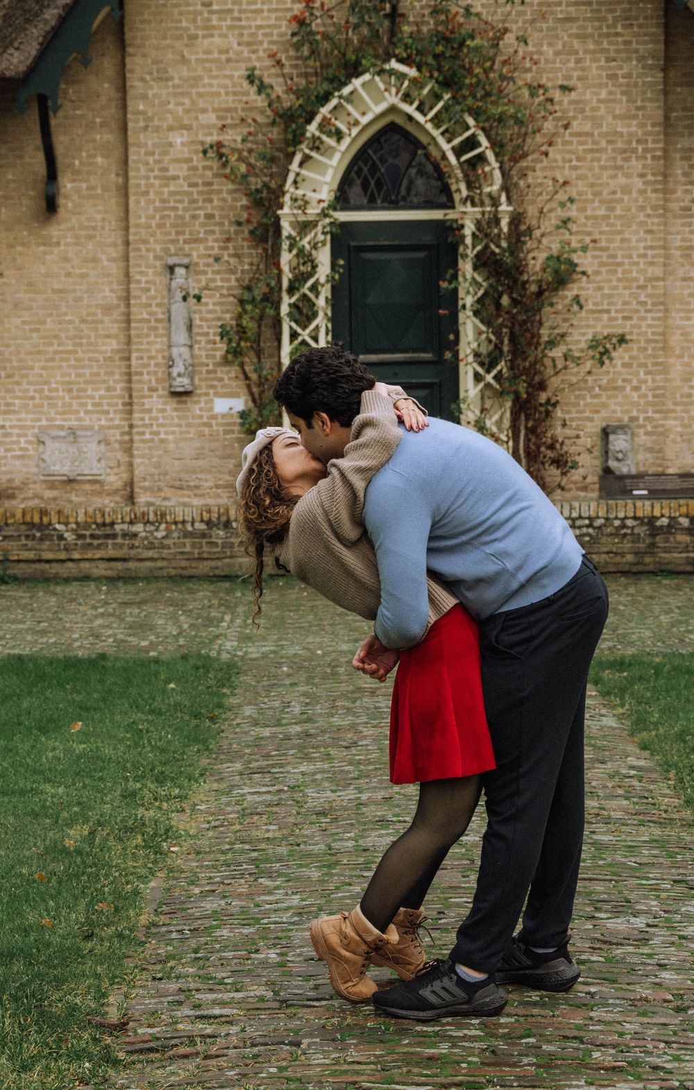 A romantic proposal scene unfolds on a cobblestone path in front of a brick building with climbing vines.