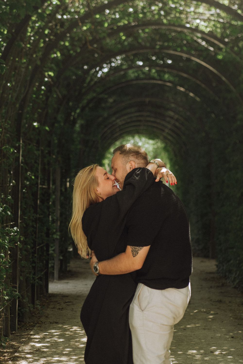 A romantic couple embraces in a lush garden tunnel with natural light filtering through the greenery.