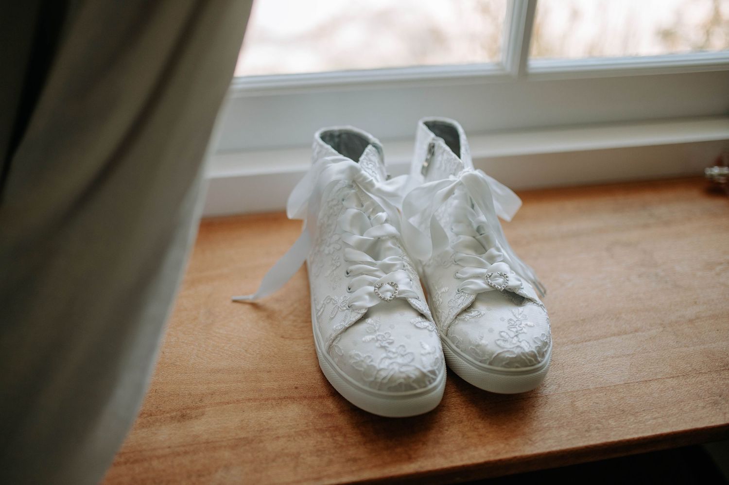 A pair of white canvas sneakers sits on a wooden window sill with natural light streaming in.