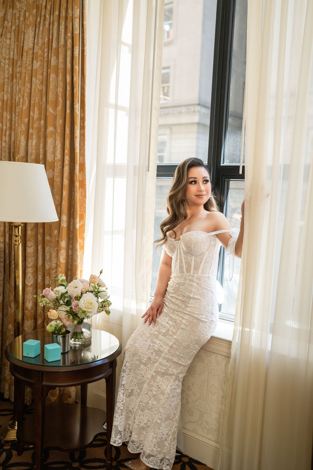 A bridal suite scene with white lace dress details and soft window light illuminating elegant floral arrangements.