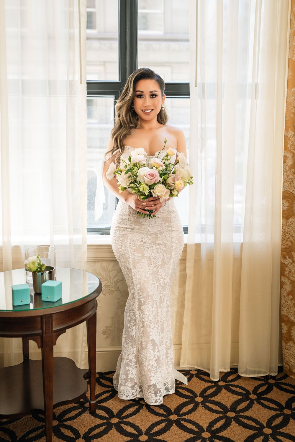 Beautiful bride in a white lace wedding gown poses elegantly in a room with patterned carpet and sheer curtains.