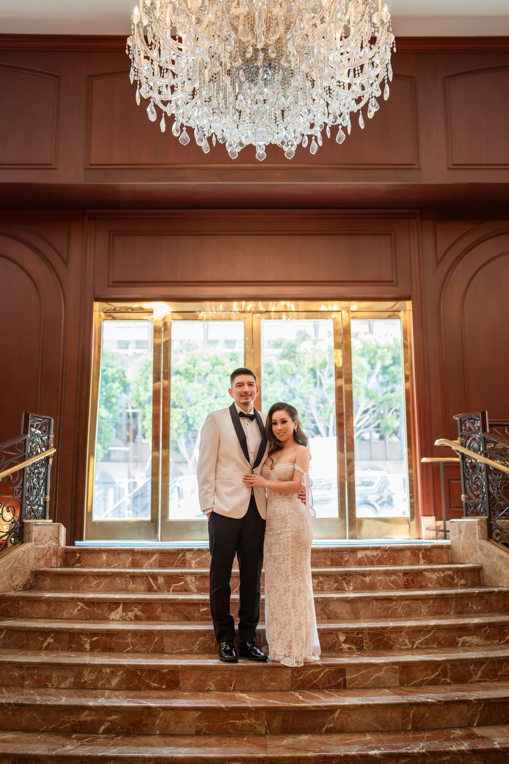 Elegant wedding couple poses beneath crystal chandeliers on marble steps in a luxurious venue.