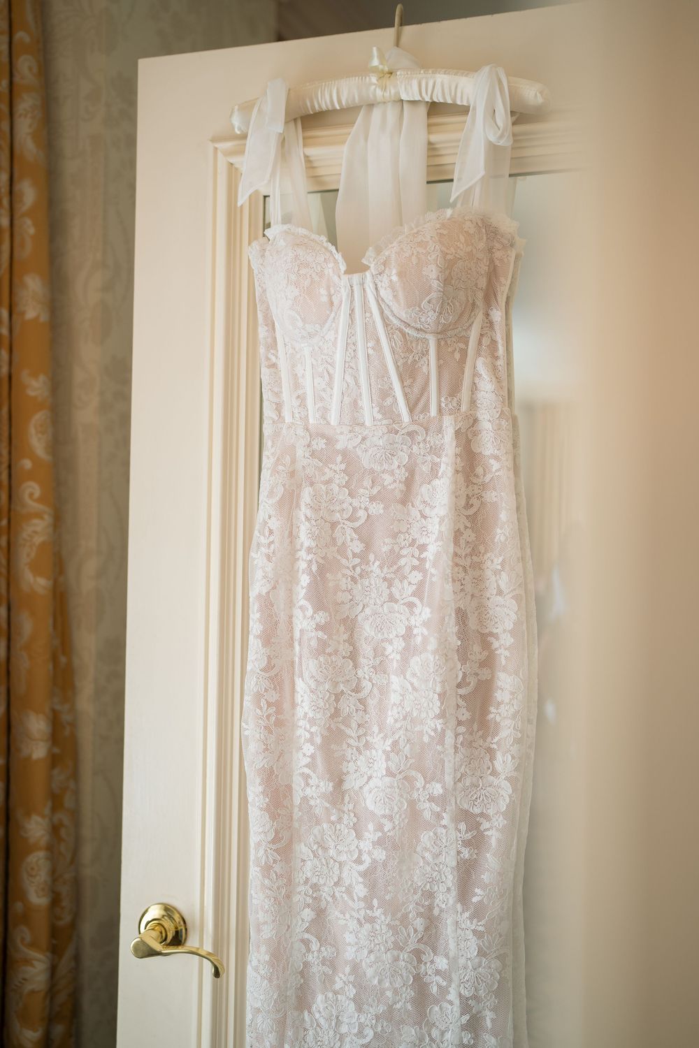 Elegant white lace wedding dress hanging on display against a cream-colored wall.