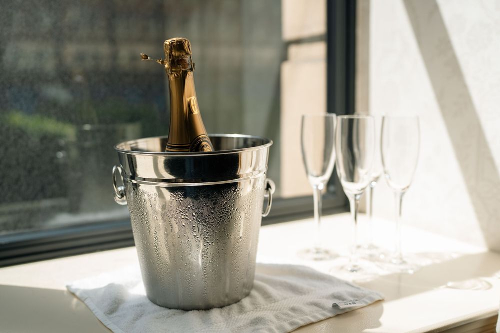 A champagne bottle chills in a frosty silver ice bucket next to empty flutes on a windowsill.