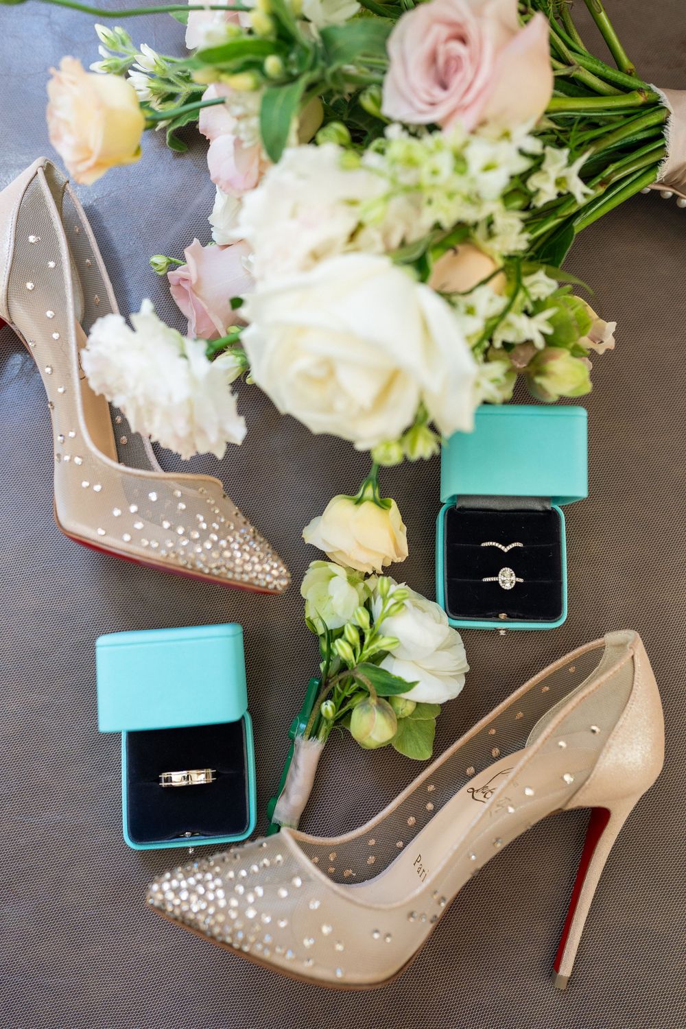 Wedding accessories including crystal-studded heels, floral bouquet and turquoise ring box arranged on grey surface.