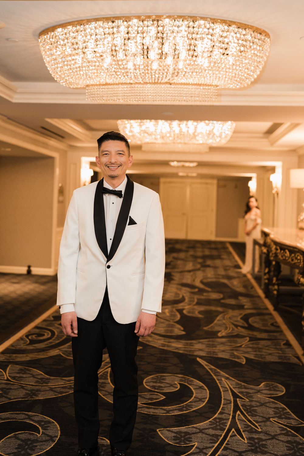 A person in a white dinner jacket and black bow tie stands in an elegant hotel hallway under crystal chandeliers.