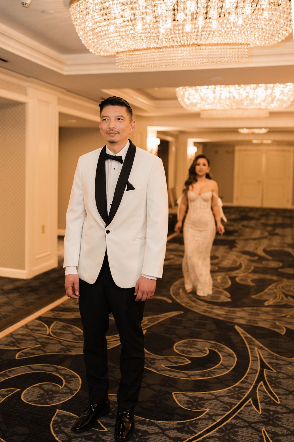 A wedding couple poses in a luxurious hotel hallway with elegant crystal chandeliers and patterned carpet.