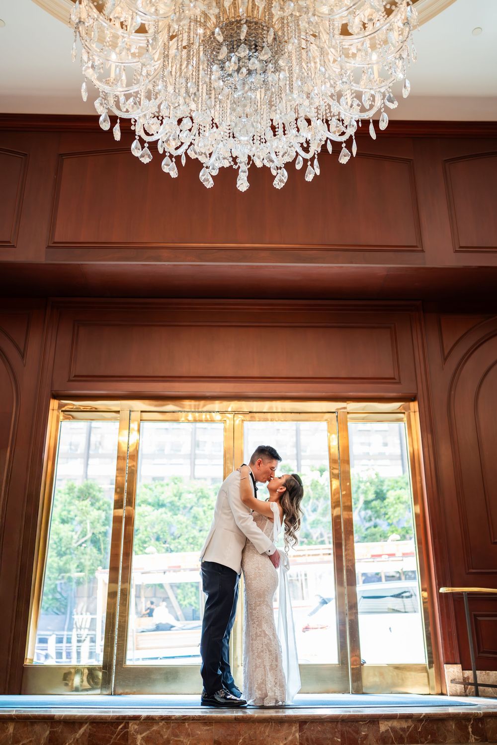 Elegant crystal chandelier hangs above a romantic scene silhouetted against bright window in an ornate room.