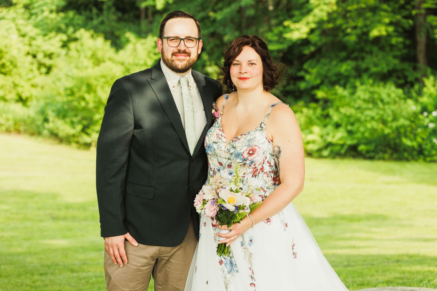 Wedding couple poses together in sunny garden setting wearing floral dress and dark suit.
