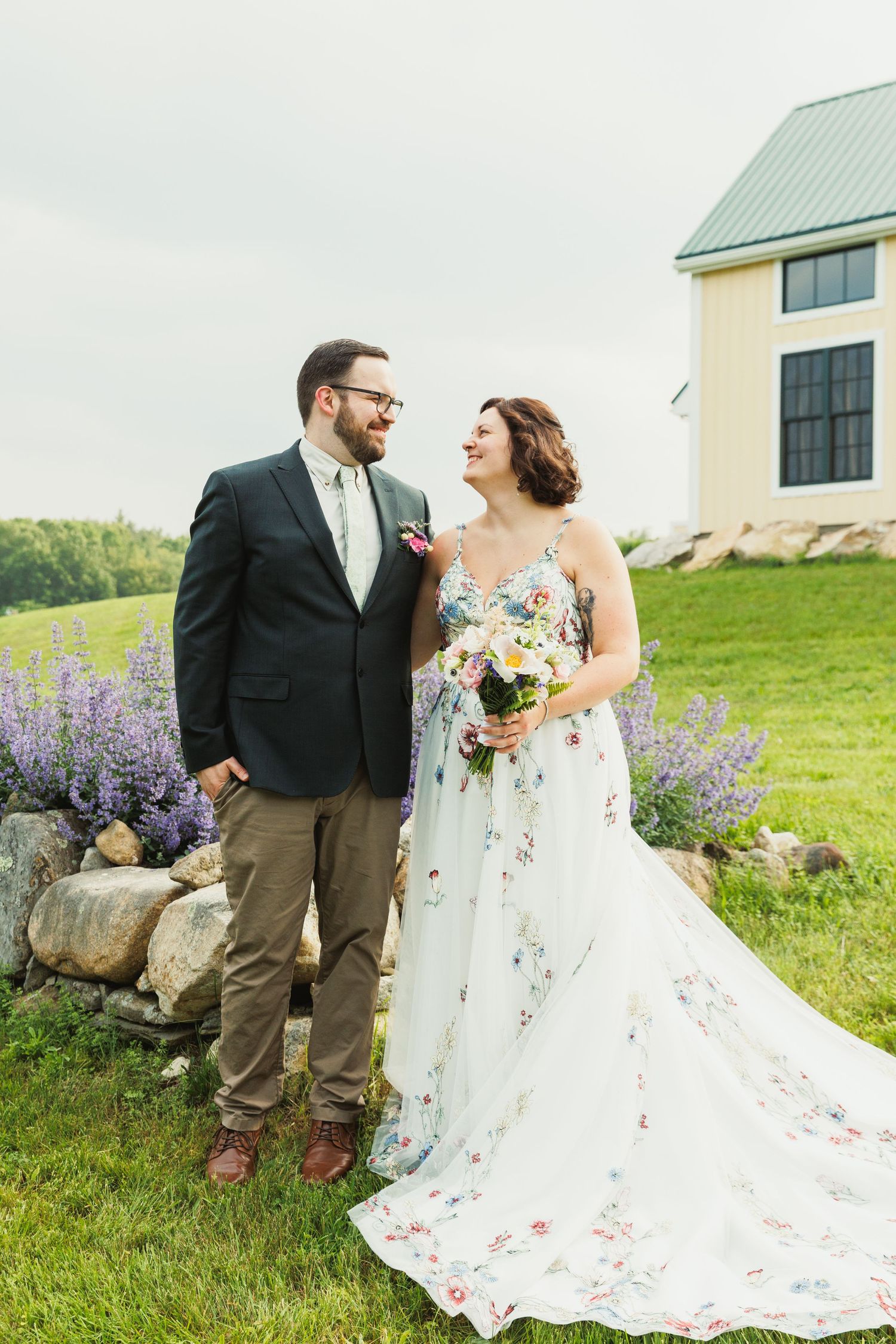 Wedding couple poses in grassy field with rustic building and purple flowers in background.