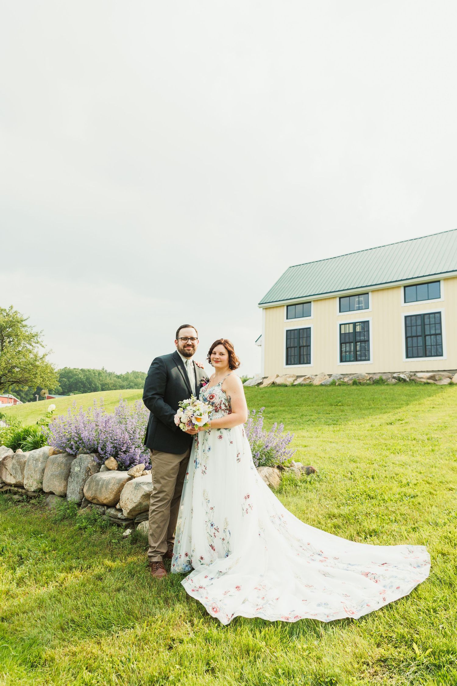 Wedding portraits in front of stone wall and lavender plants with cream colored building in background.