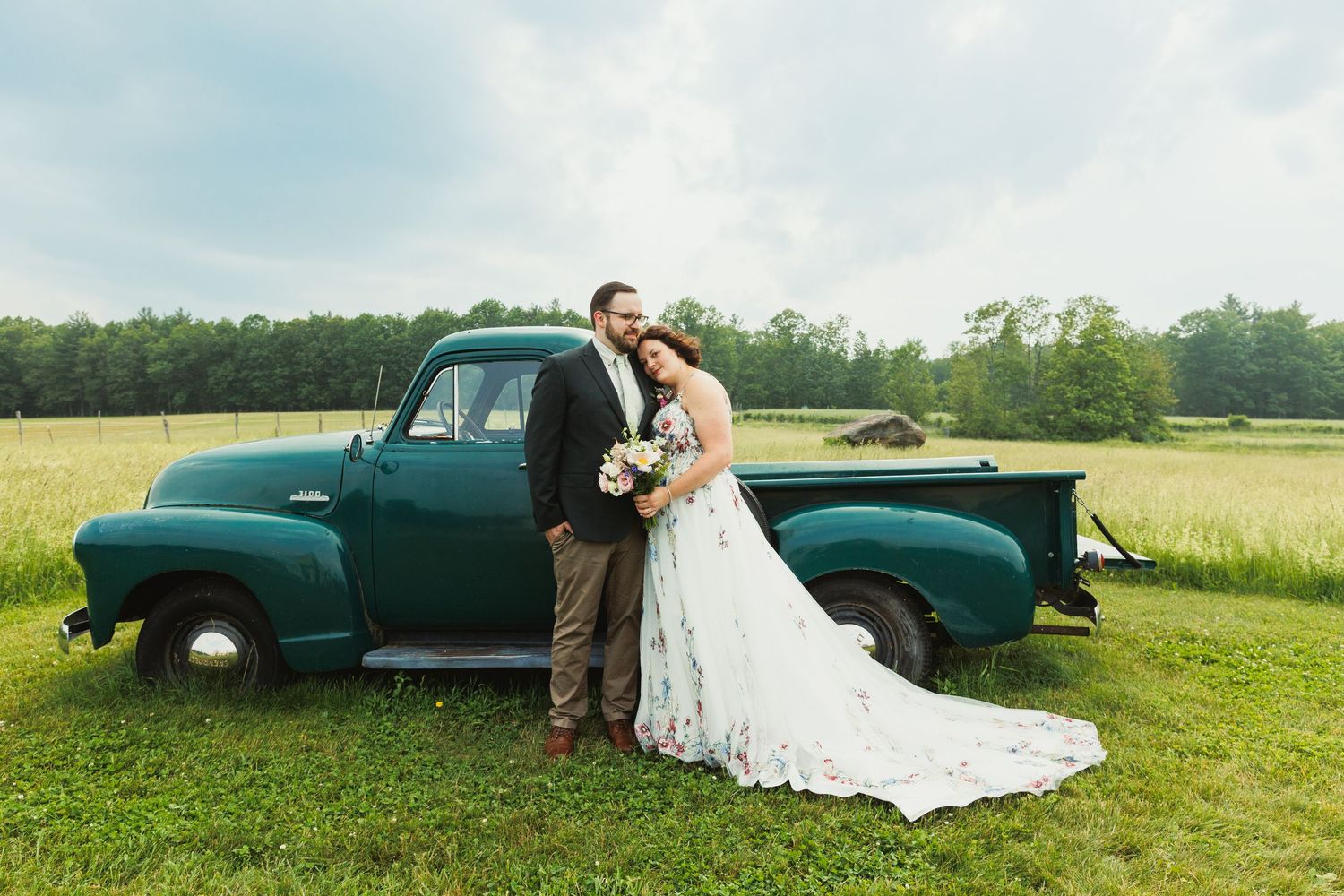 A couple poses together near a vintage teal pickup truck in a grassy field.