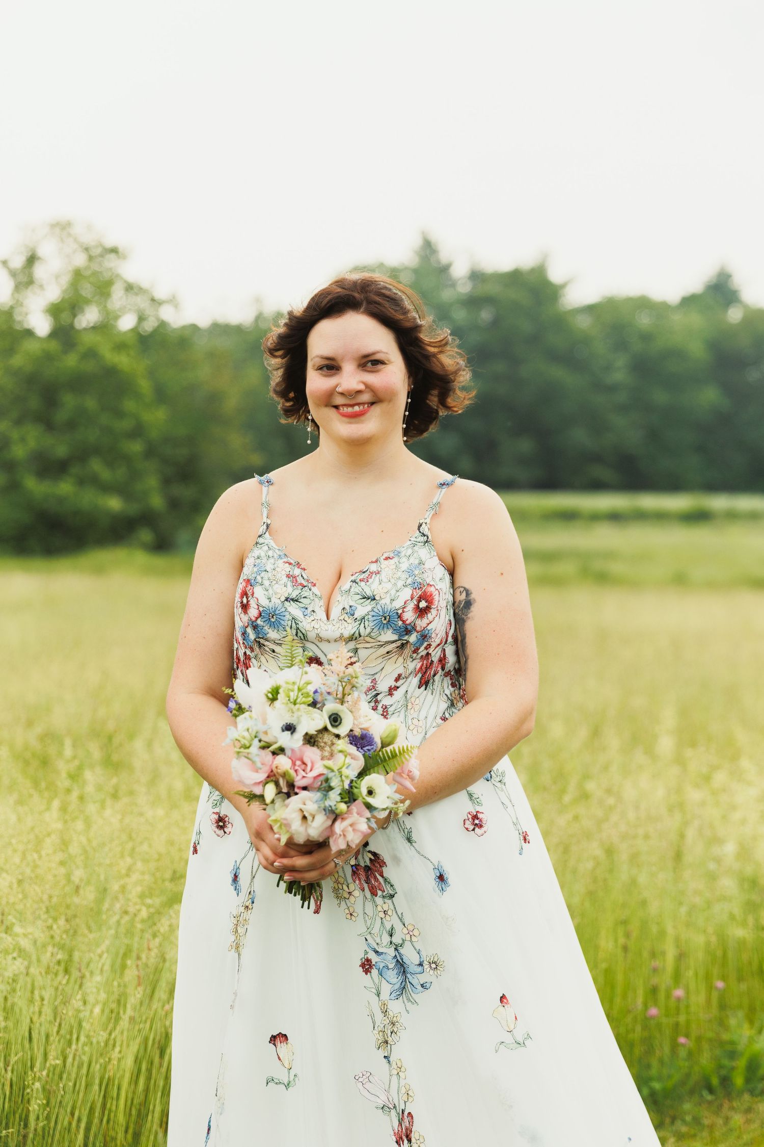 A bride in a floral wedding dress holds a colorful bouquet in a summer meadow.