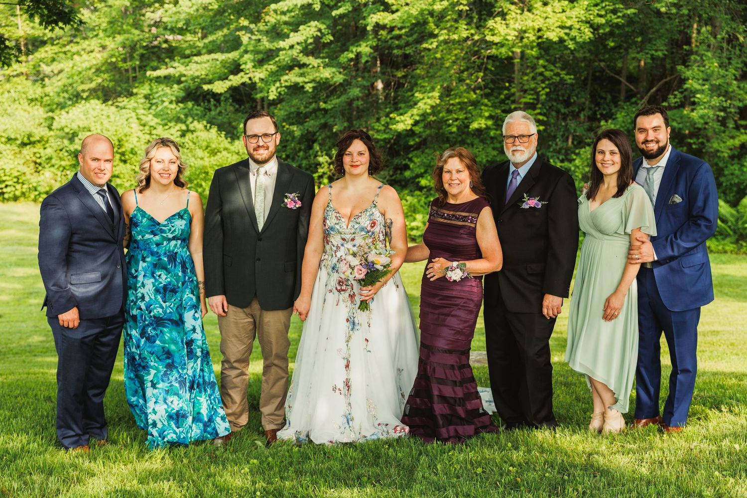 Wedding party members pose together on grass with trees in background wearing coordinated blue and burgundy attire.