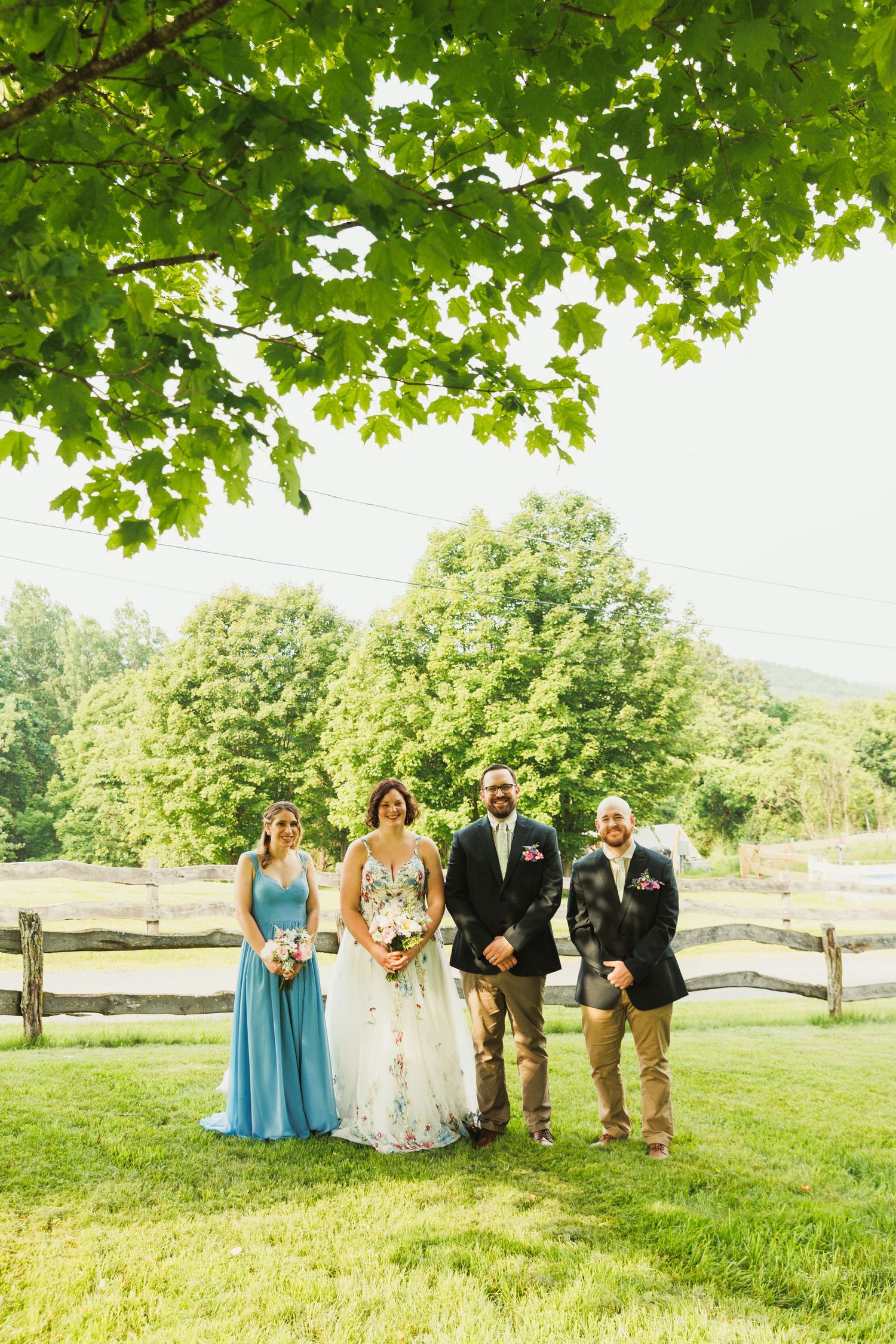 Wedding party stands under green tree canopy in outdoor garden setting with natural lighting.
