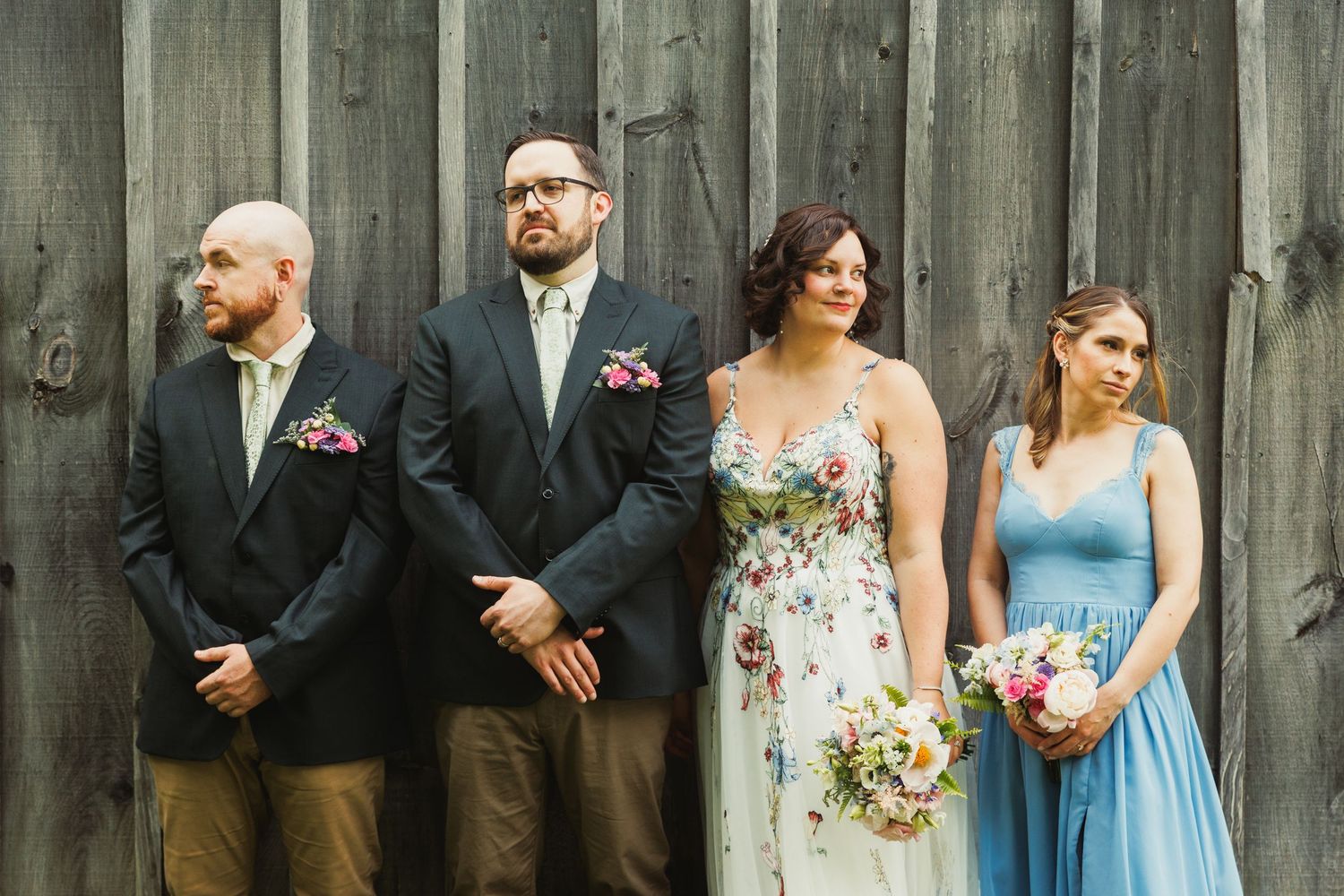 Wedding party poses against rustic wooden barn wall wearing mix of formal and semi-formal attire.