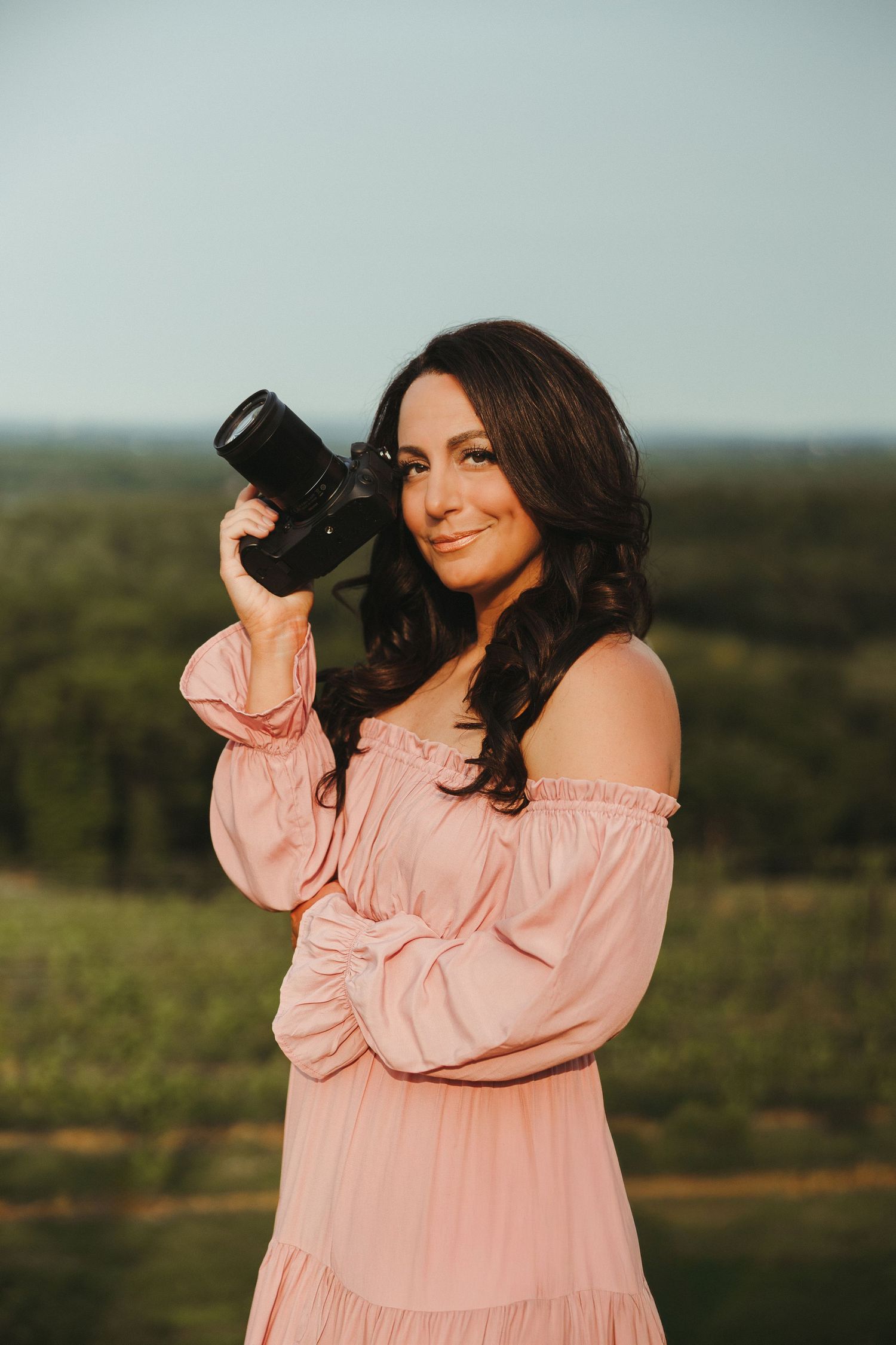 Woman in flowing pink dress poses with professional camera against scenic landscape.