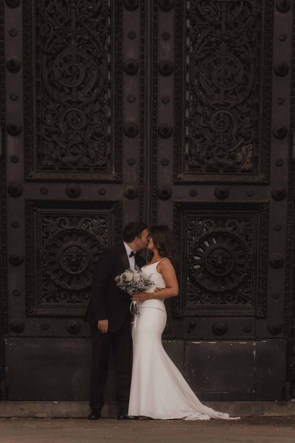 Wedding party poses against ornate dark doors with elegant architectural details in moody lighting.