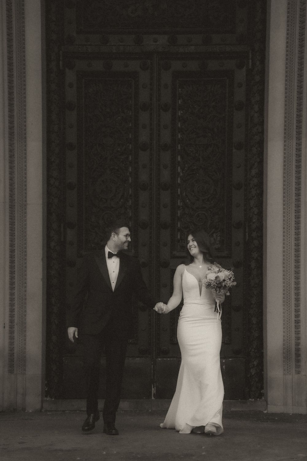 A black and white photograph shows people walking in a formal setting with tall columns and elegant architecture.