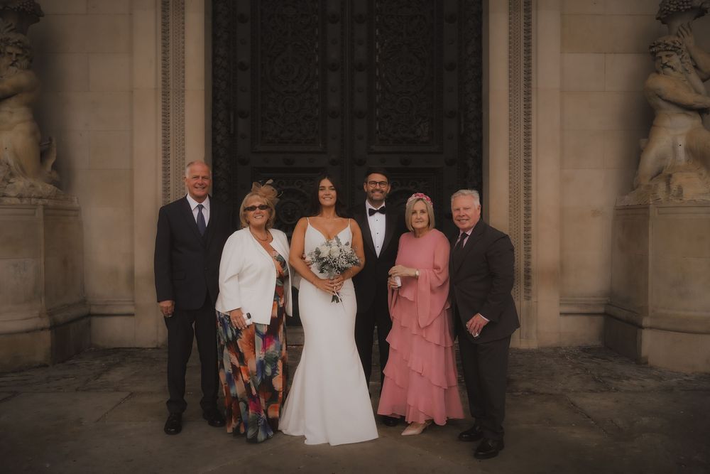 A group of wedding attendees pose together in a formal setting with stone columns and architecture in the background.
