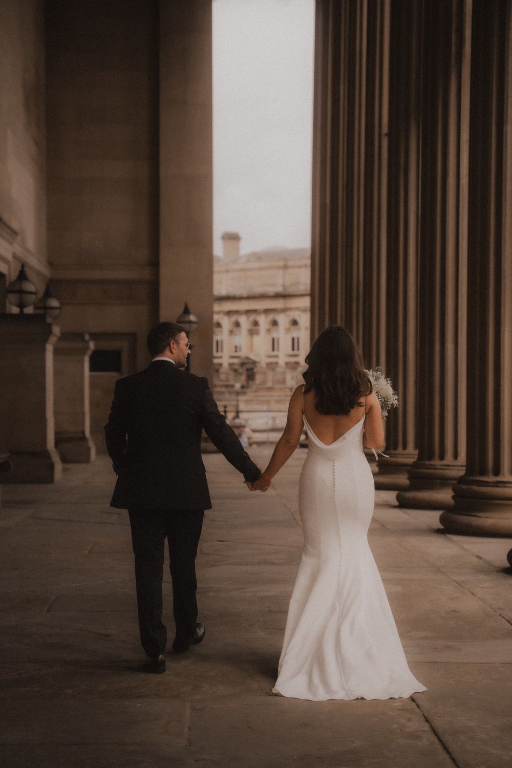 A couple walks hand in hand through a grand colonnade with tall stone pillars in a romantic wedding setting.