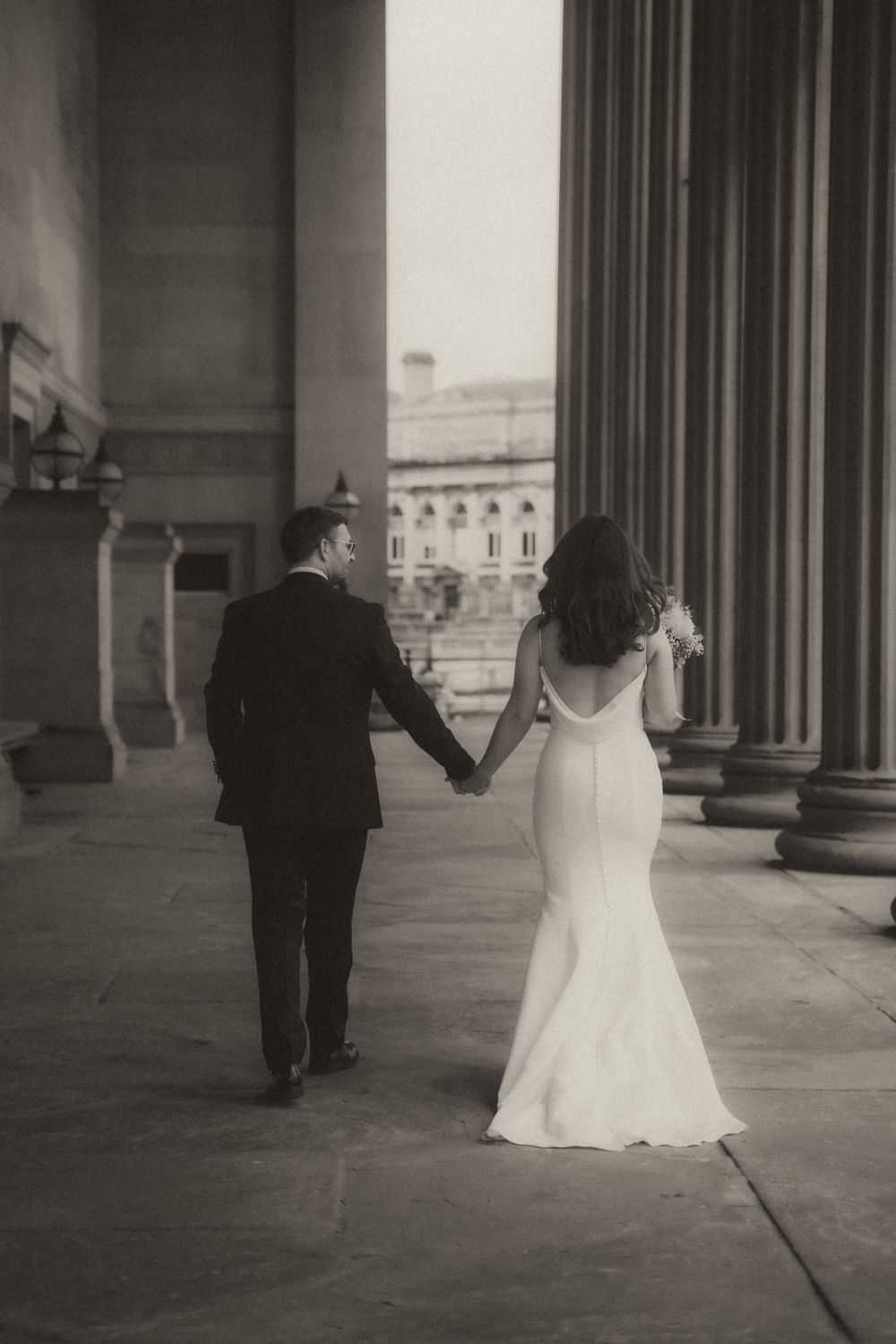 A black and white wedding photo showing a couple walking hand in hand between classical columns of a grand building.