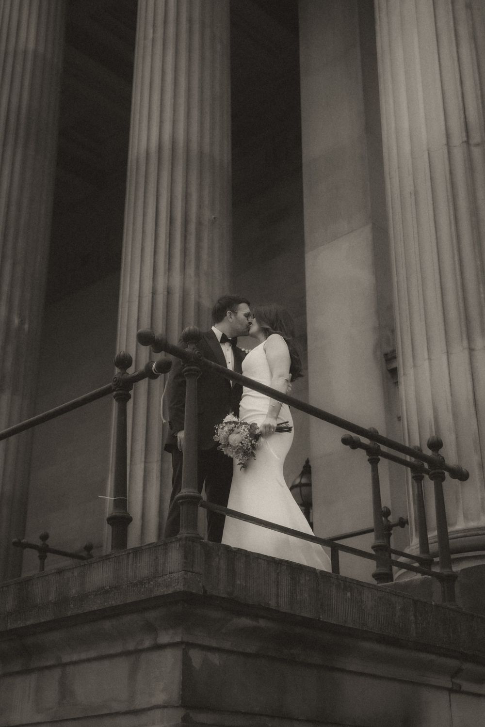 A romantic wedding couple shares a kiss on a balcony surrounded by classical stone columns at dusk.